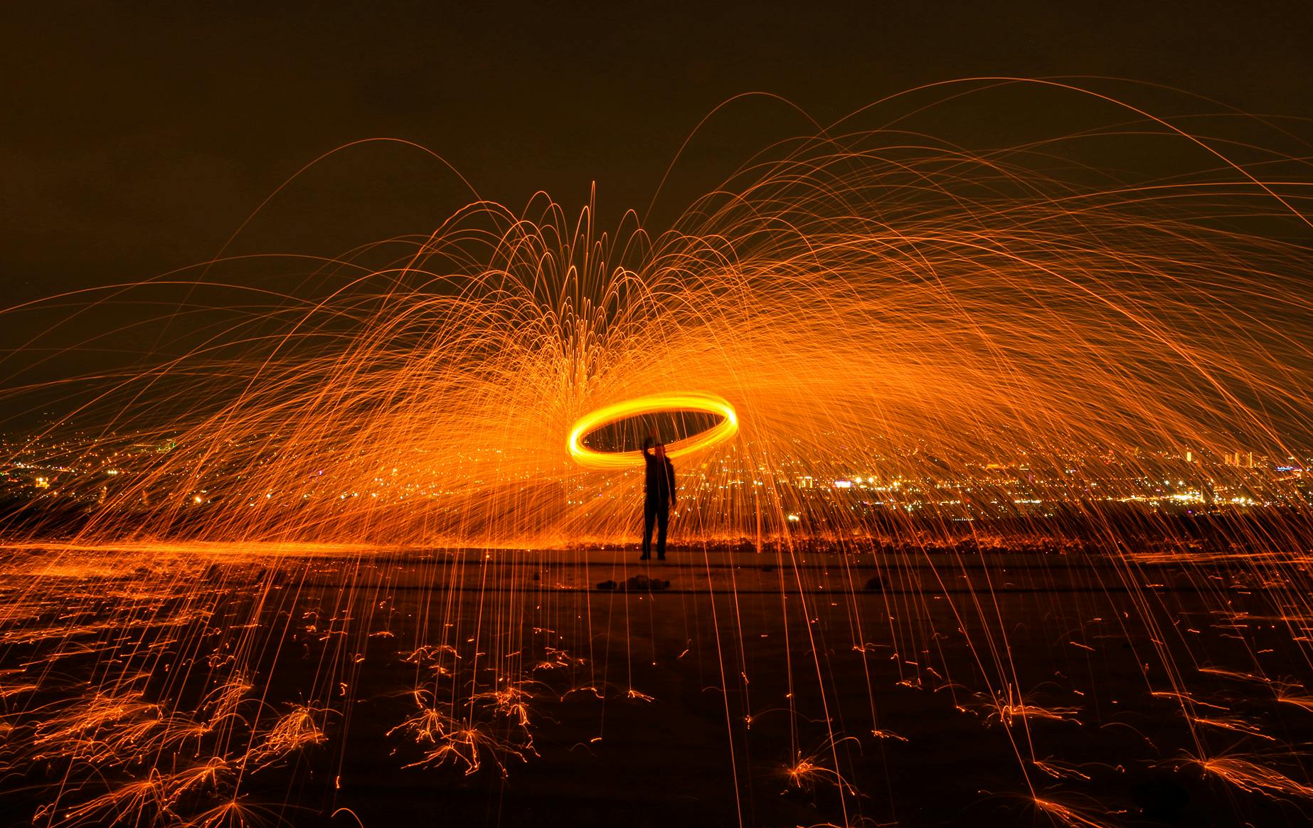 A long-exposure photograph of a person silhouetted against a dark sky, holding spinning steel wool that creates a halo of bright orange light and sparks, with city lights visible in the background.