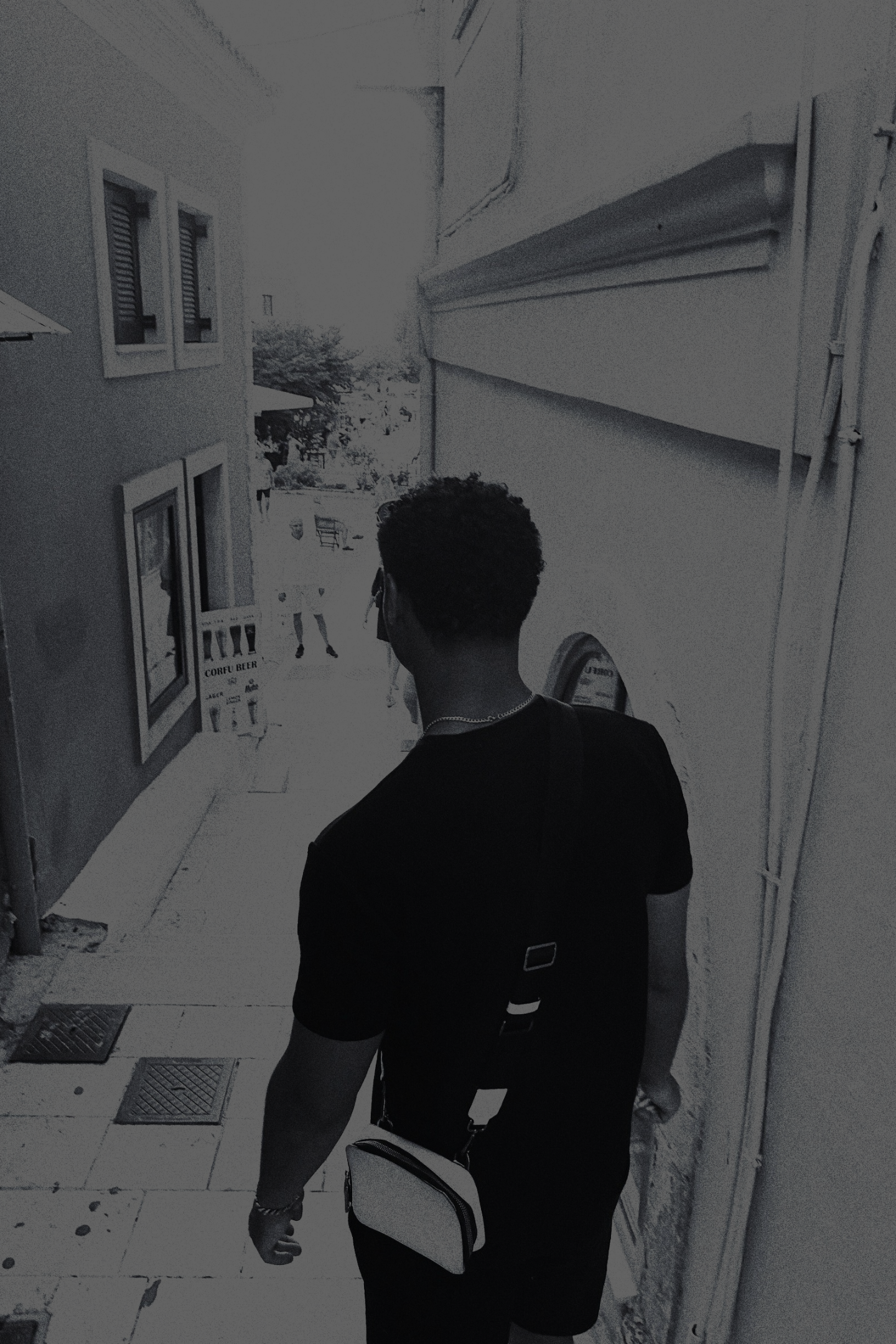 A grayscale image of a man with his back to the viewer, wearing a black shirt and shorts, walking down a narrow street with buildings and a glimpse of greenery in the background.